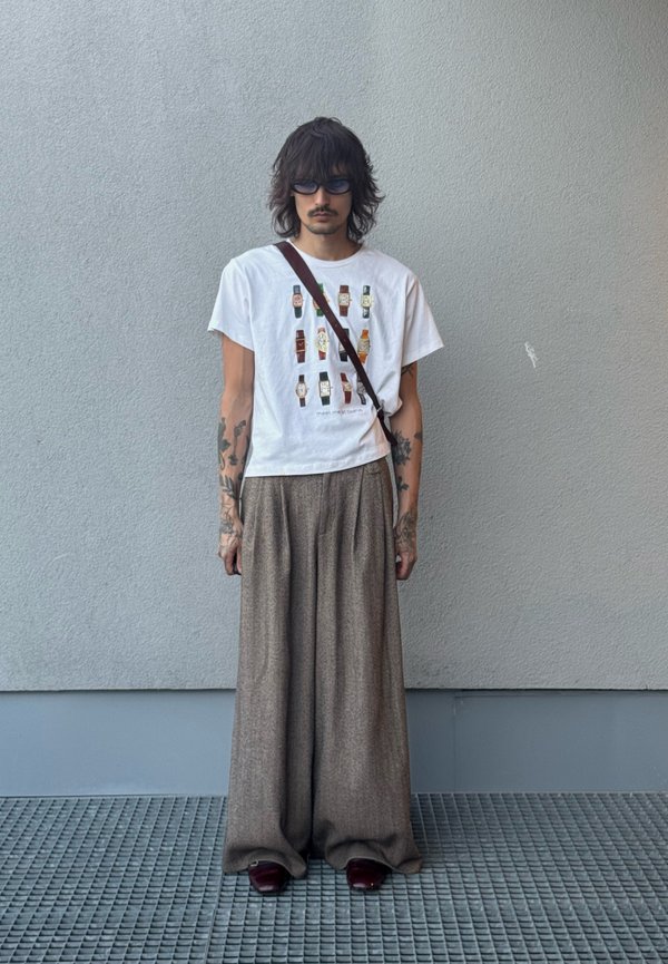 Personne aux cheveux foncés mi-longs et tatouages portant des lunettes de soleil, un t-shirt blanc avec des motifs de montres, un pantalon marron large et des chaussures foncées, debout contre un mur.
