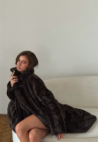 Mørkebrun pelsfrakke i kunstig materiale med høj krave og tekstureret mønster, draperet over en lysefarvet sofa, der viser blødt, plyssede materiale.