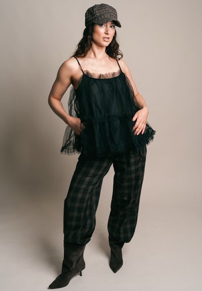 Mujer con una blusa negra sin mangas translúcida, pantalones de cuadros, botas negras y una gorra tipo newsboy de cuadros, de pie con las manos en los bolsillos.