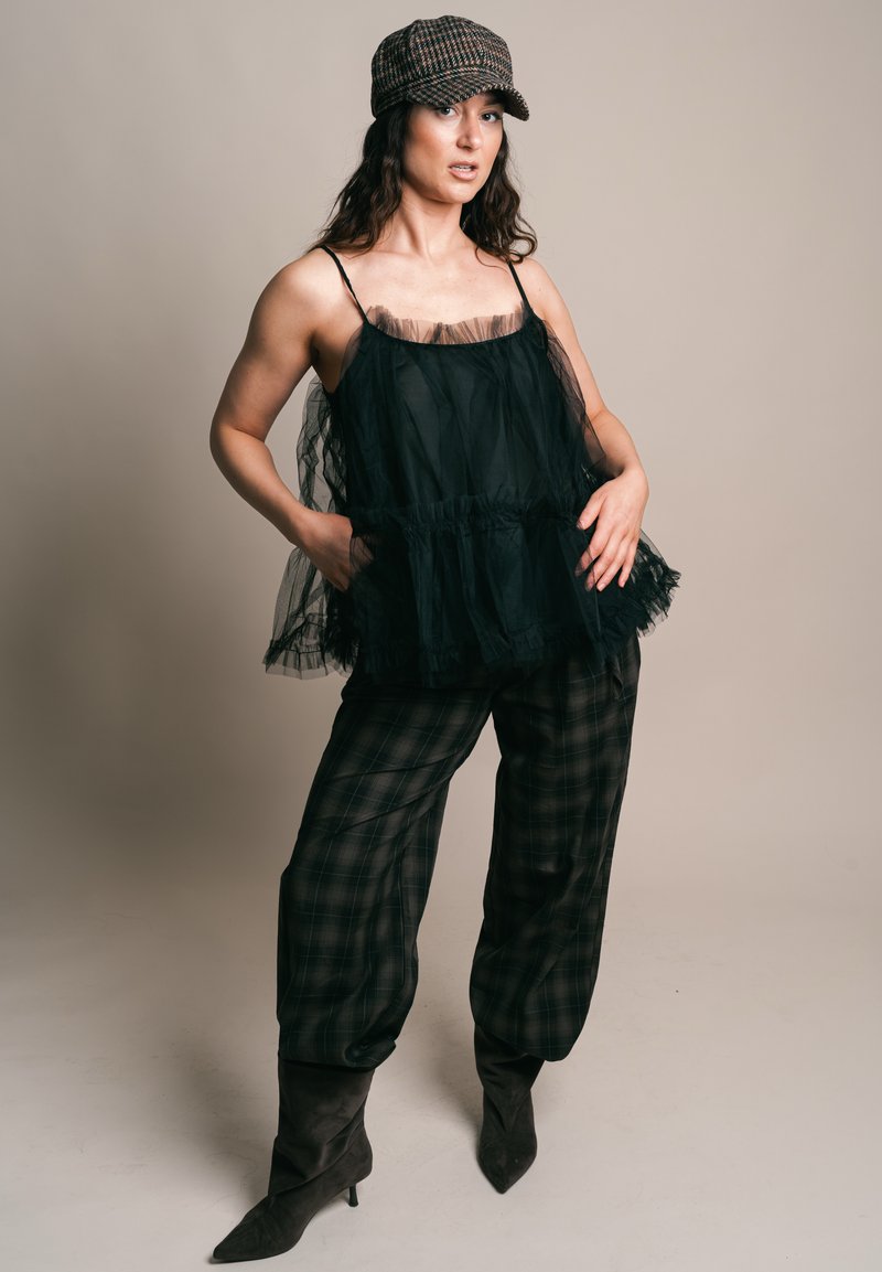 Nő fekete, áttetsző ujjatlan felsőt, kockás nadrágot, fekete csizmát és kockás newsboy sapkát visel, és zsebében tartja a kezét.