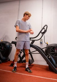Tricou sportiv cu dungi gri și pantaloni scurți gri deschis, purtat cu pantofi negri și șosete albe. Fundalul prezintă echipamente de sală pe un podea roșie.