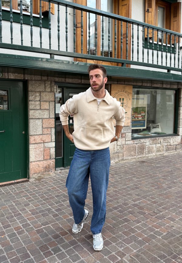 Beige zip-up tröja med hög krage och framficka; bärs med lösa blå jeans och grå sportskor på en kullerstensgata.