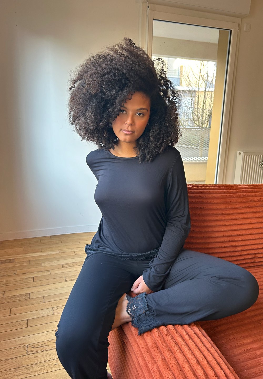 Top nero a maniche lunghe e pantaloni con dettagli in pizzo, seduta su un divano arancione con texture. Pavimento in legno naturale e finestra sullo sfondo.