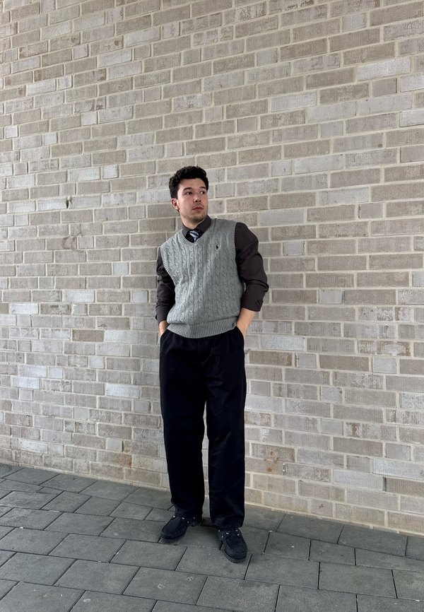 Jeune homme portant un gilet en maille gris par-dessus une chemise foncée et une cravate, pantalon et chaussures noirs, debout les mains dans les poches devant un mur de briques gris.