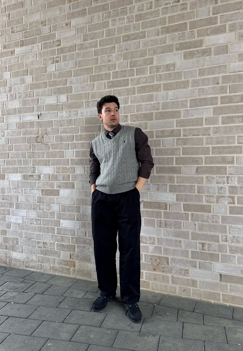 Jeune homme portant un gilet en tricot gris sur une chemise foncée et une cravate, pantalon et chaussures noirs, debout avec les mains dans les poches devant un mur de briques gris.