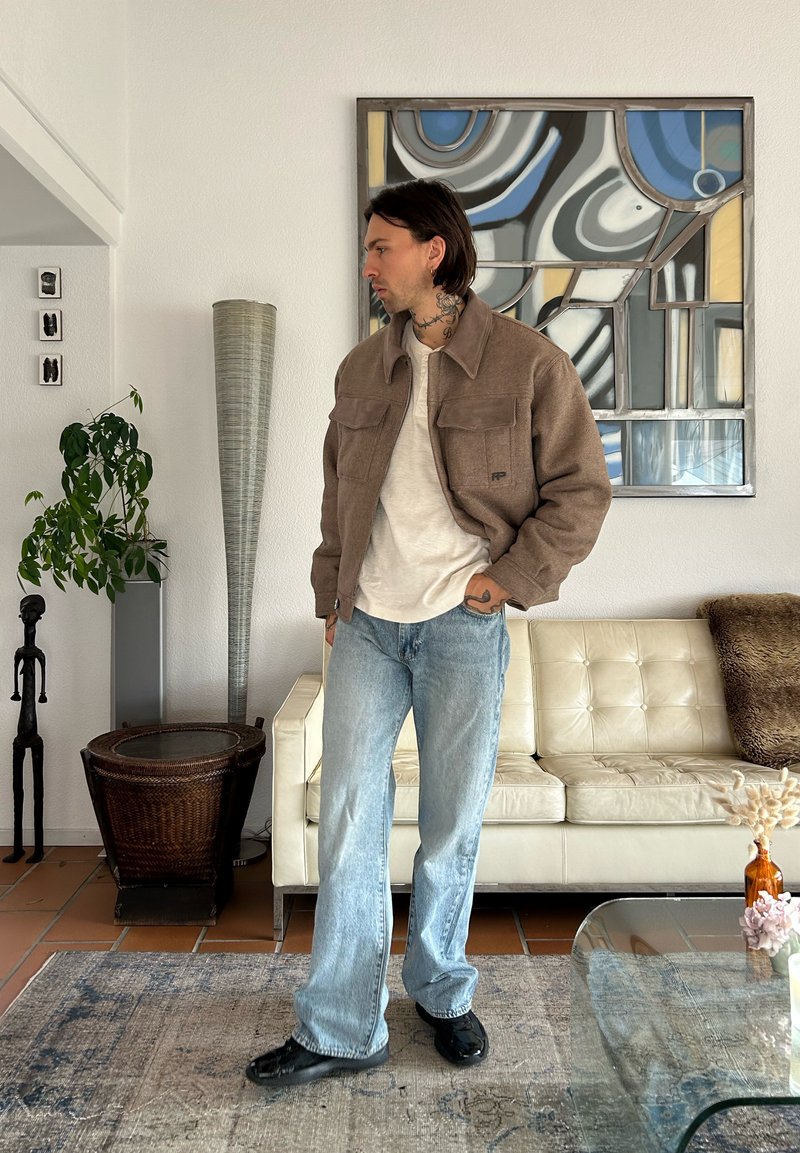 Veste marron texturée avec deux poches avant, chemise beige, jean bleu clair et chaussures noires. Intérieur minimaliste avec des plantes et des œuvres d'art.