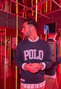 Mornarska trenirka z besedilom "POLO" v svetlo modri barvi, nameščena preko bele srajce z ozkimi črtami. Ozadje vsebuje rdeče neonske luči in ogledala.