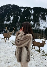 Plašč iz umetnega krzna različnih odtenkov rjave in sive, z mehko teksturo. Snega pokrajina z jelenom v ozadju.