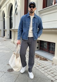 Jeansjacka, krämfärgad tröja, grå byxor, vita sneakers och en stor beige tote-väska. Scenen visar en stenlagd trottoar och arkitektoniska detaljer.