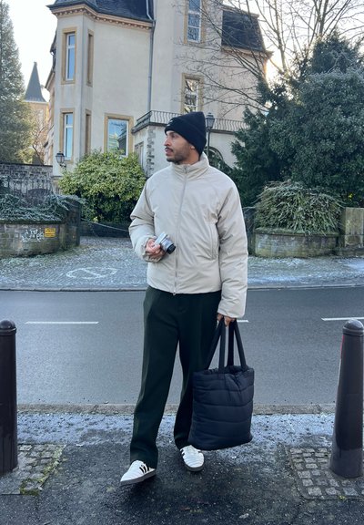 Chaqueta de plumas en beige, gorro negro, pantalones verde oscuro, zapatillas blancas, sosteniendo una cámara y llevando una bolsa tote negra. Fondo urbano.
