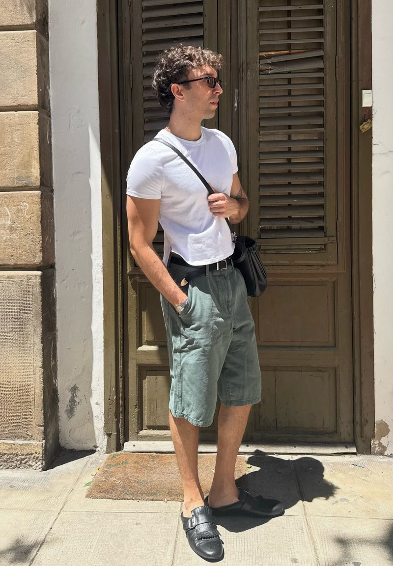 Homme portant des lunettes de soleil, un t-shirt blanc, un short vert et des chaussures noires, se tient devant une porte en bois marron avec une main dans la poche.