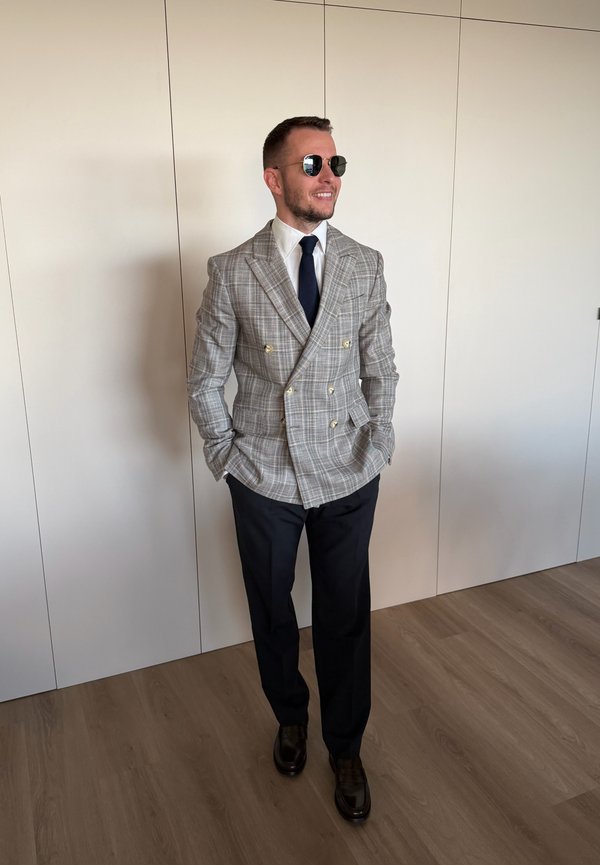 Hombre con blazer de cuadros gris cruzado, camisa blanca, corbata oscura, pantalones negros y gafas de sol, de pie con las manos en los bolsillos sobre suelo de madera.