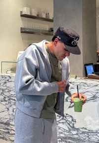 Szary sweatshirt i pasujące spodnie na zielonej koszulce. Trzyma zielony napój z czarną słomką. W tle marmurowy blat.
