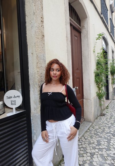 Top peplum negro con tirantes blancos, combinado con pantalones blancos con cordón. Cabello rizado, de pie junto a un letrero de tienda y una pared de piedra texturizada.