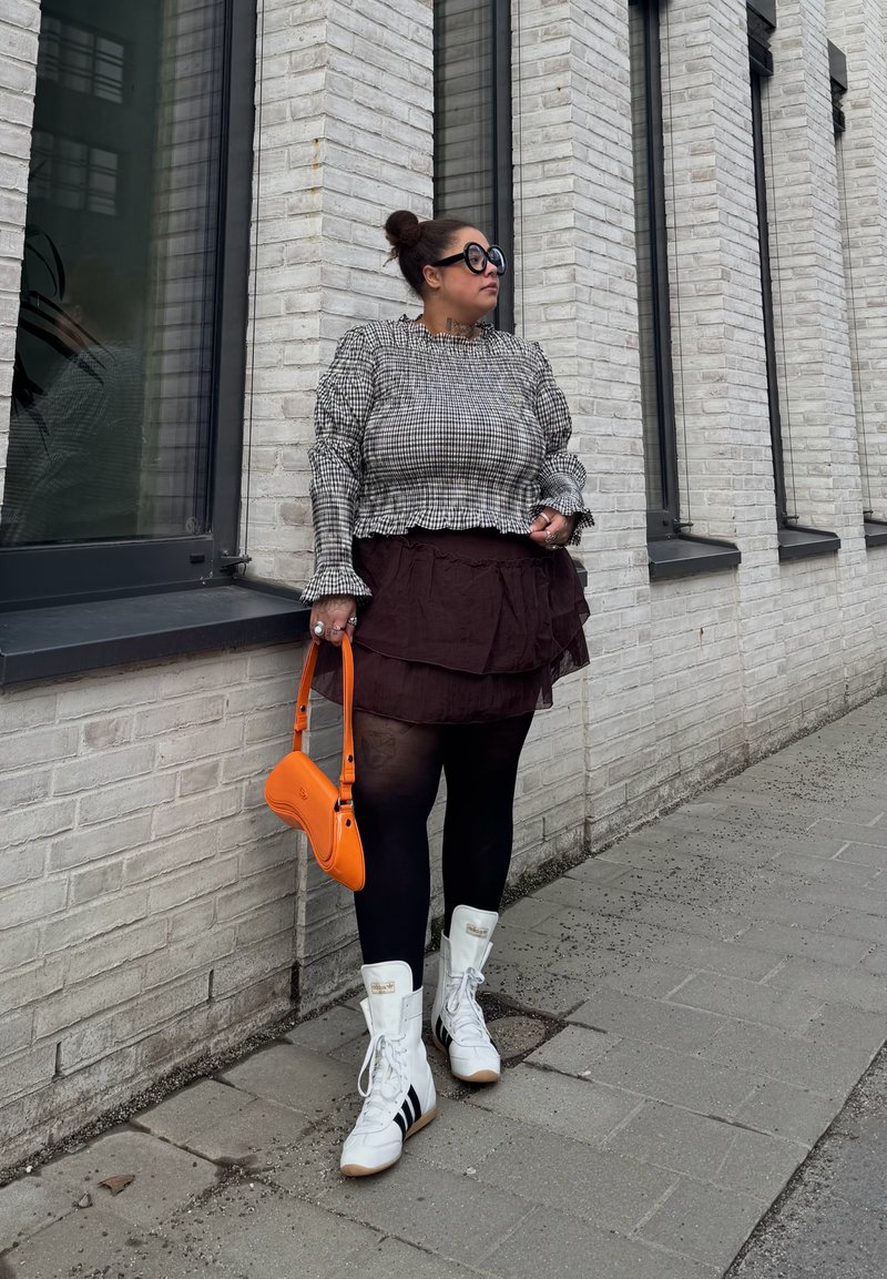 Person in a checkered top, layered black skirt, black tights, white Adidas high-top sneakers, and large round sunglasses, holding an orange handbag.