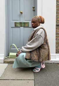 Mesterséges szőrme mellény barna árnyalatokkal, szürke, túlméretezett pulóver fölött. Világoskék pliszírozott szoknya, rózsaszín szoborszerű saruk és halványzöld oldaltáska.