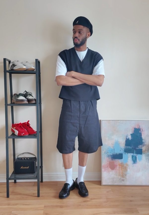 Uomo con basco nero, gilet nero, camicia bianca, pantaloncini neri, calzini bianchi e scarpe nere in piedi con le braccia incrociate vicino a uno scaffale per scarpe e a un dipinto astratto.