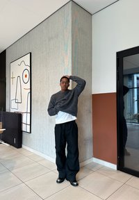 Homem com óculos, suéter cinza, camisa branca e calças pretas está em pé dentro de casa contra uma parede de canto texturizada, perto de uma obra de arte abstrata e uma porta de vidro.