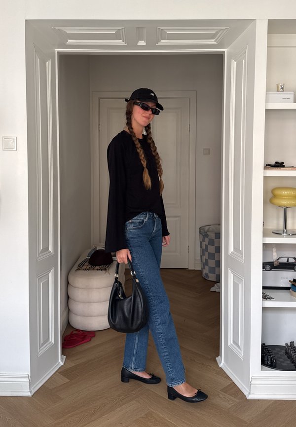 Mujer con mangas largas negras, vaqueros, zapatos negros y gorra, sosteniendo un bolso negro, de pie en una puerta blanca en el interior.