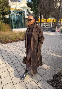 Femme portant un long manteau marron, un pull, une jupe et des bottes noires marchant sur un chemin pavé près de plantations et d'une structure en verre dans une zone urbaine ensoleillée.