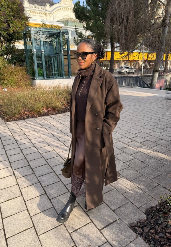 Nő barna hosszú kabátban, pulóverben, szoknyában és fekete csizmában, aki egy napos városi területen, növények és üvegépület közelében, burkolt úton sétál.