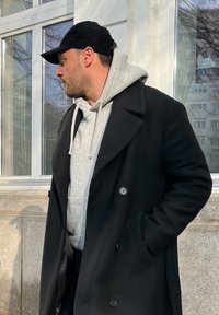 Svart dubbelknäppt rock över grå hoodie, bärs med svart keps. Står bredvid ett glasfönster med en texturerad vägg i bakgrunden.