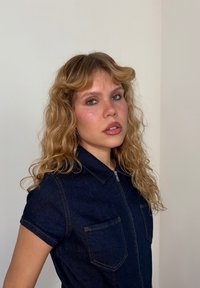 Denim-haalarilla, jossa on kaulus, lyhyet hihat, kolme etutaskua, etuvetoketju ja kontrastoiva oranssi ommel. Kihara vaaleanpunainen hius.