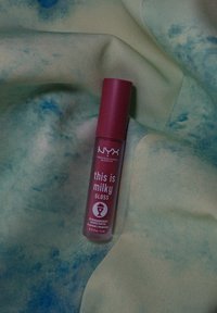 Η NYX Professional Makeup milky gloss σε ροζ σωλήνα πάνω από μια ελαφριά μπλε-πράσινη υφασμάτινη επιφάνεια με υφή. Η ετικέτα περιλαμβάνει λεπτομέρειες για τη γεύση και τον όγκο.