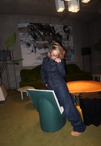 Τζιν ολόσωμο φόρεμα με εφαρμοστή σχεδίαση, ξεθωριασμένο μπλε χρώμα και αντιθετικές ραφές. Χαλαρή καθιστική διάταξη στο φόντο και στρογγυλό ξύλινο τραπέζι.