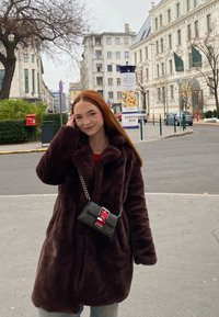 Καφέ, χνουδωτό υπερμεγέθες παλτό με μεγάλο γιακά, φορεμένο πάνω από μια κόκκινη μπλούζα. Μια μαύρη τσάντα ώμου με πολύχρωμη λεπτομέρεια κρέμεται από τον ώμο.