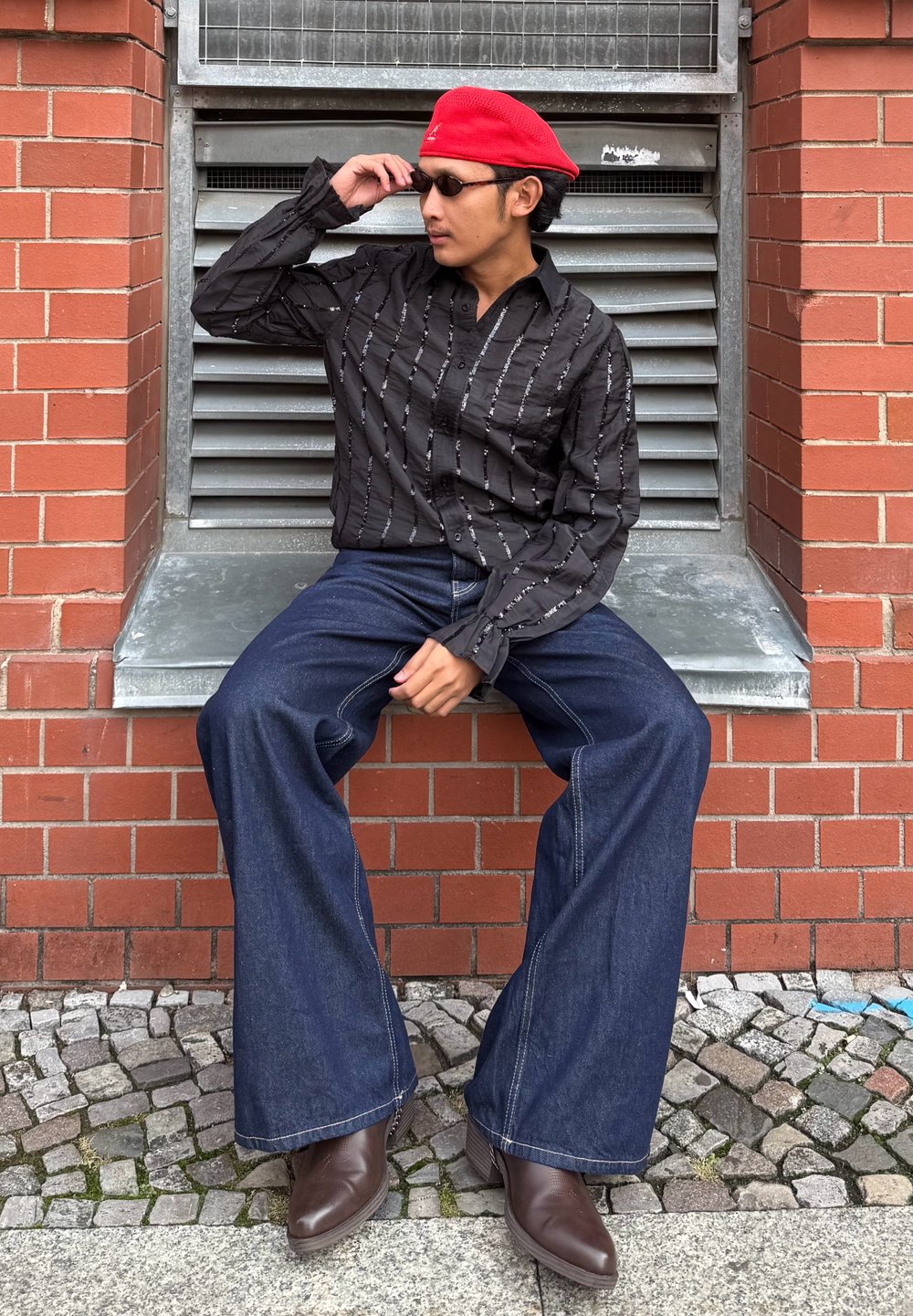 Čierna košeľa na zapínanie s bielymi vertikálnymi pruhmi, rozšírené tmavomodré džínsy a hnedé kožené čižmy. Červený baret a slnečné okuliare ako doplnky.