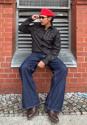 Camisa negra abotonada con rayas verticales blancas, vaqueros azul oscuro acampanados y botas de cuero marrón. Boina roja y gafas de sol como accesorios.