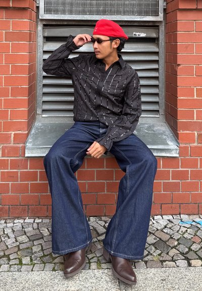 Camisa negra abotonada con rayas verticales blancas, vaqueros azul oscuro acampanados y botas de cuero marrón. Boina roja y gafas de sol como accesorios.