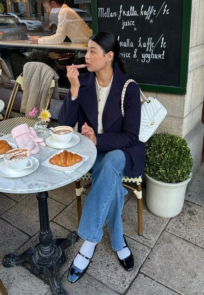 Mujer sentada en una mesa de café con croissants y café, aplicándose bálsamo labial, vistiendo un abrigo azul marino y jeans, junto a un menú de pizarra.