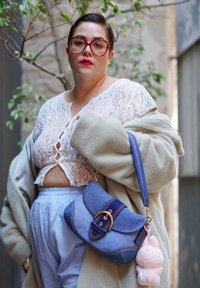 Mala de ombro em denim com ferragens douradas, detalhes azuis e um charm em pelúcia rosa. O modelo veste uma blusa de rendas e um casaco oversized, combinado com calças claras.