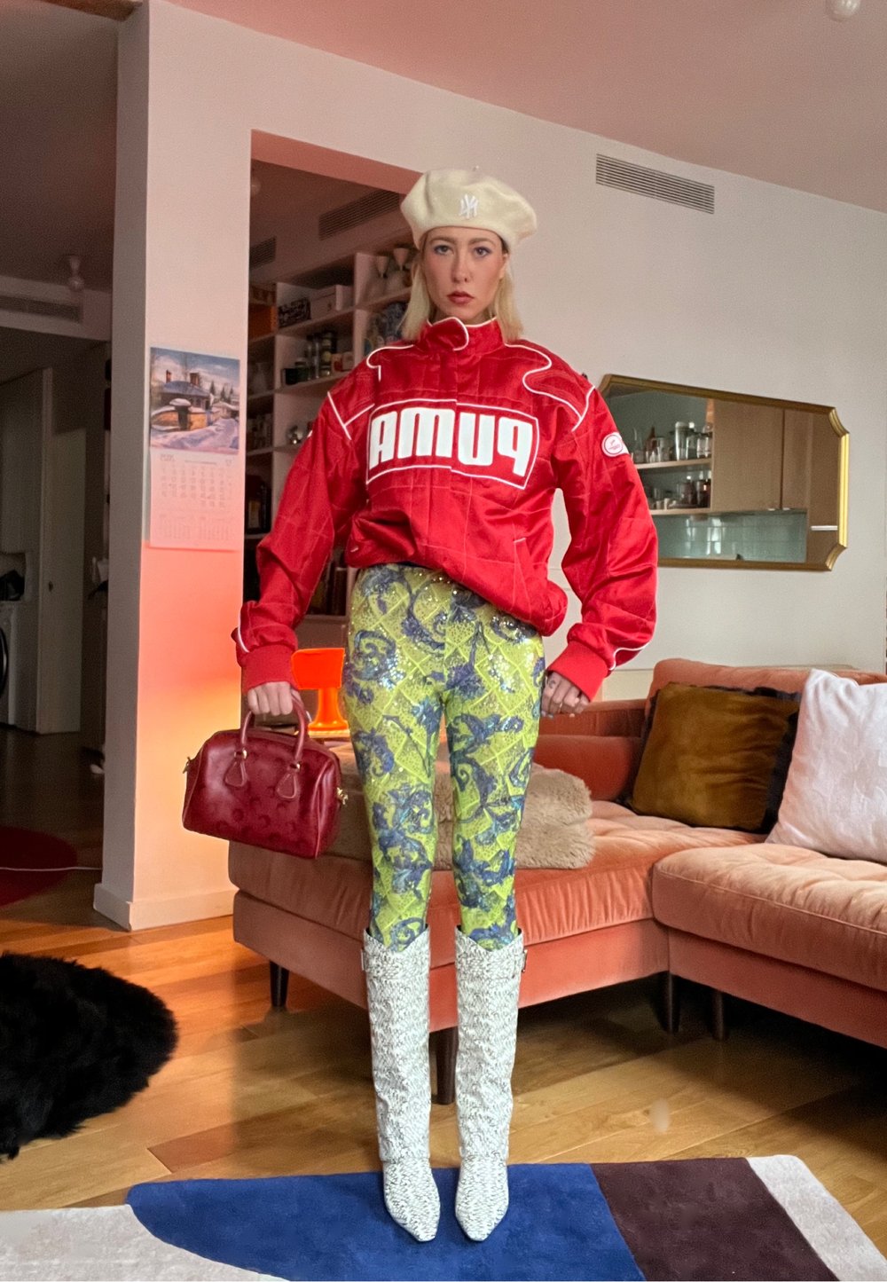 Femme portant une veste rouge Puma, un pantalon à motifs jaune-vert, de hautes bottes blanches et un béret blanc, tenant un sac à main rouge dans un salon.