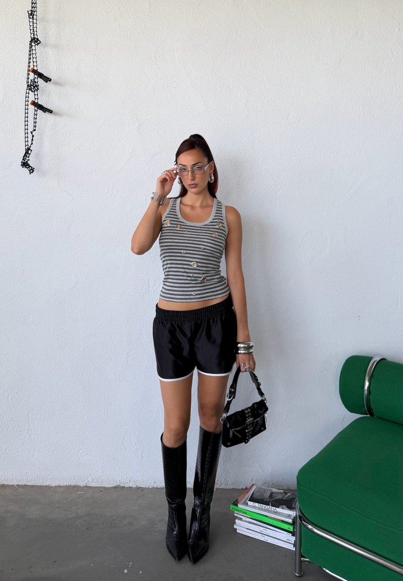 Jeune femme portant un débardeur rayé, un short noir et des bottes jusqu'aux genoux, debout près d'une chaise verte tenant un sac à main noir à l'intérieur.