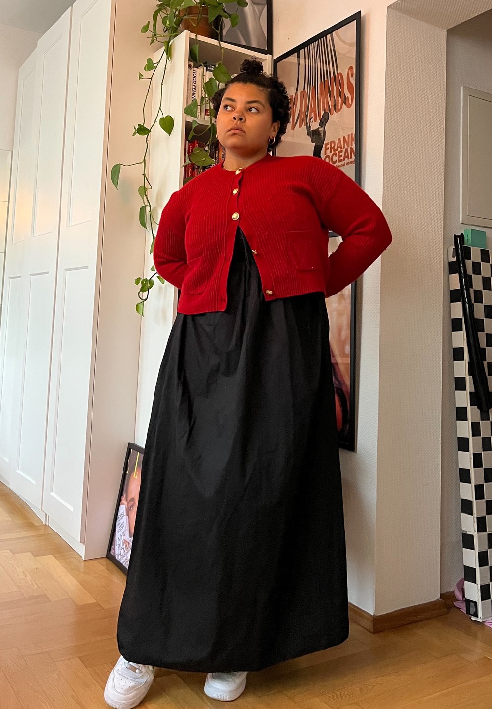 Κόκκινο ριγέ κασκόλ με χρυσά κουμπιά πάνω από μια ροή μαύρη μακριά φόρεμα, συνδυασμένο με άσπρα αθλητικά παπούτσια, σε ένα καλά φωτισμένο εσωτερικό χώρο.