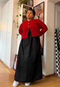 Κόκκινο ριγέ κασκόλ με χρυσά κουμπιά πάνω από μια ροή μαύρη μακριά φόρεμα, συνδυασμένο με άσπρα αθλητικά παπούτσια, σε ένα καλά φωτισμένο εσωτερικό χώρο.