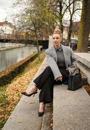 Blazer gris oversize, blusa oscura, pantalones negros de pierna ancha y tacones puntiagudos negros. Bolso negro con diseño estructurado a un lado. Hojas de fondo.