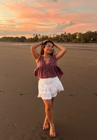 Femme en haut violet sans manches et jupe blanche, debout pieds nus sur une plage de sable au coucher du soleil, avec des palmiers et des bâtiments en arrière-plan.