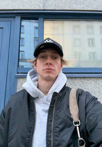 Zwarte bomberjack over een grijze hoodie, met een zwarte pet met een logo. Lichtbruine schouderriem met zichtbare metalen onderdelen. Stedelijke achtergrond.