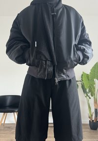 Schwarze Bomberjacke mit einem gerippten schwarzen Schal, Reißverschluss und elastischen Bündchen, kombiniert mit schwarzen Shorts mit weitem Bein. Glatte Textur.