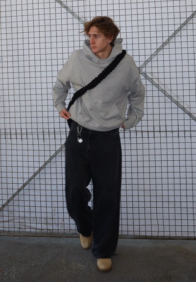 Sudadera gris con capucha y bolsillo frontal, pantalones anchos negros, zuecos beiges y un bolso negro de strap trenzado. El fondo tiene un patrón de rejilla.