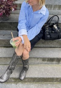Camisa listrada azul clara e branca, saia preta, botas cinzentas até ao joelho com fivelas, segurando um copo transparente com um straws, sentada nos degraus.