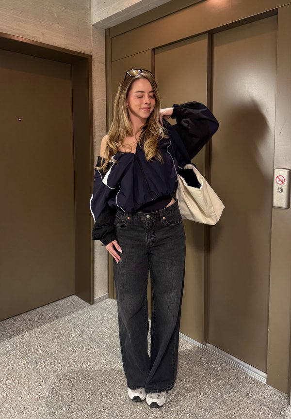 Giovane donna con capelli lunghi, che indossa una giacca nera scollata, jeans scuri, scarpe da ginnastica bianche e porta una borsa bianca, in piedi vicino alle porte dell’ascensore.