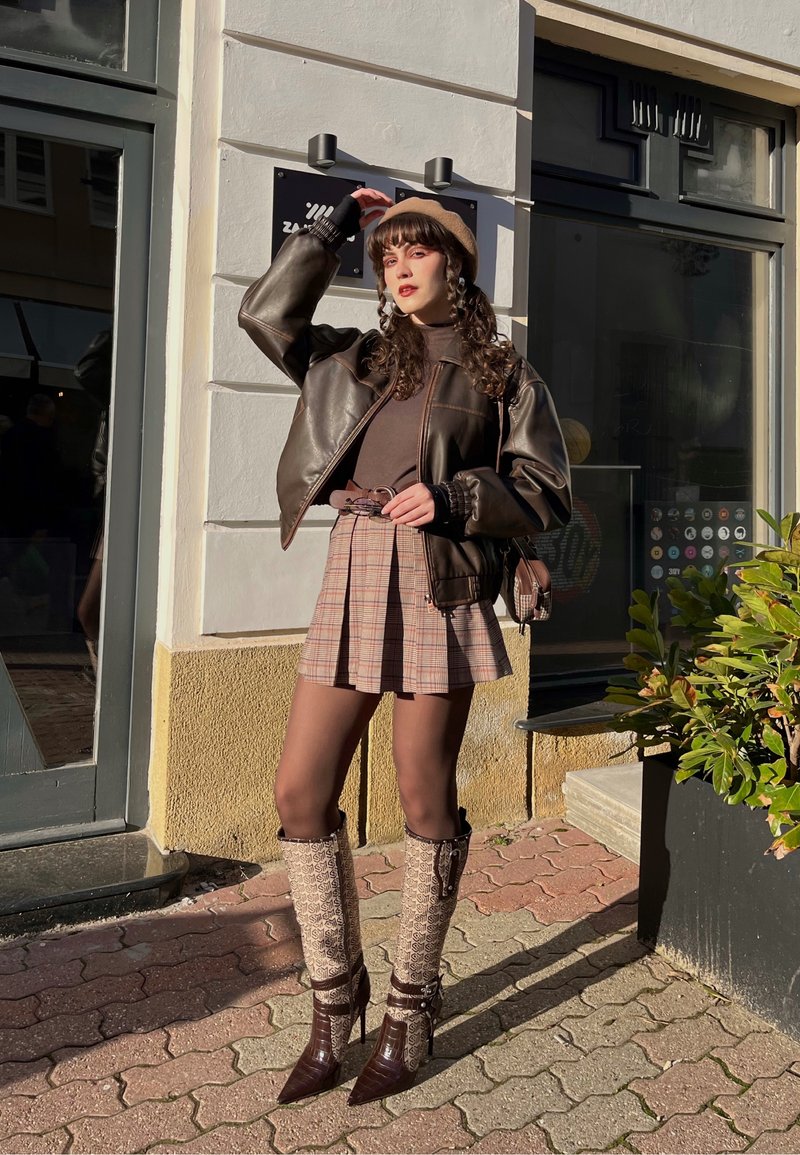 Jeune femme en veste en cuir marron, jupe à carreaux, bottes beige et béret posant devant un bâtiment ensoleillé près de plantes.
