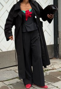 Άτομο που φοράει μαύρο παλτό τύπου τρεντς, μαύρο παντελόνι με φαρδιά μπατζάκια, μαύρο και πράσινο τοπ με κόκκινη λεπτομέρεια λουλουδιού, κόκκινα παπούτσια, κρατώντας μια μαύρη τσάντα χειρός.