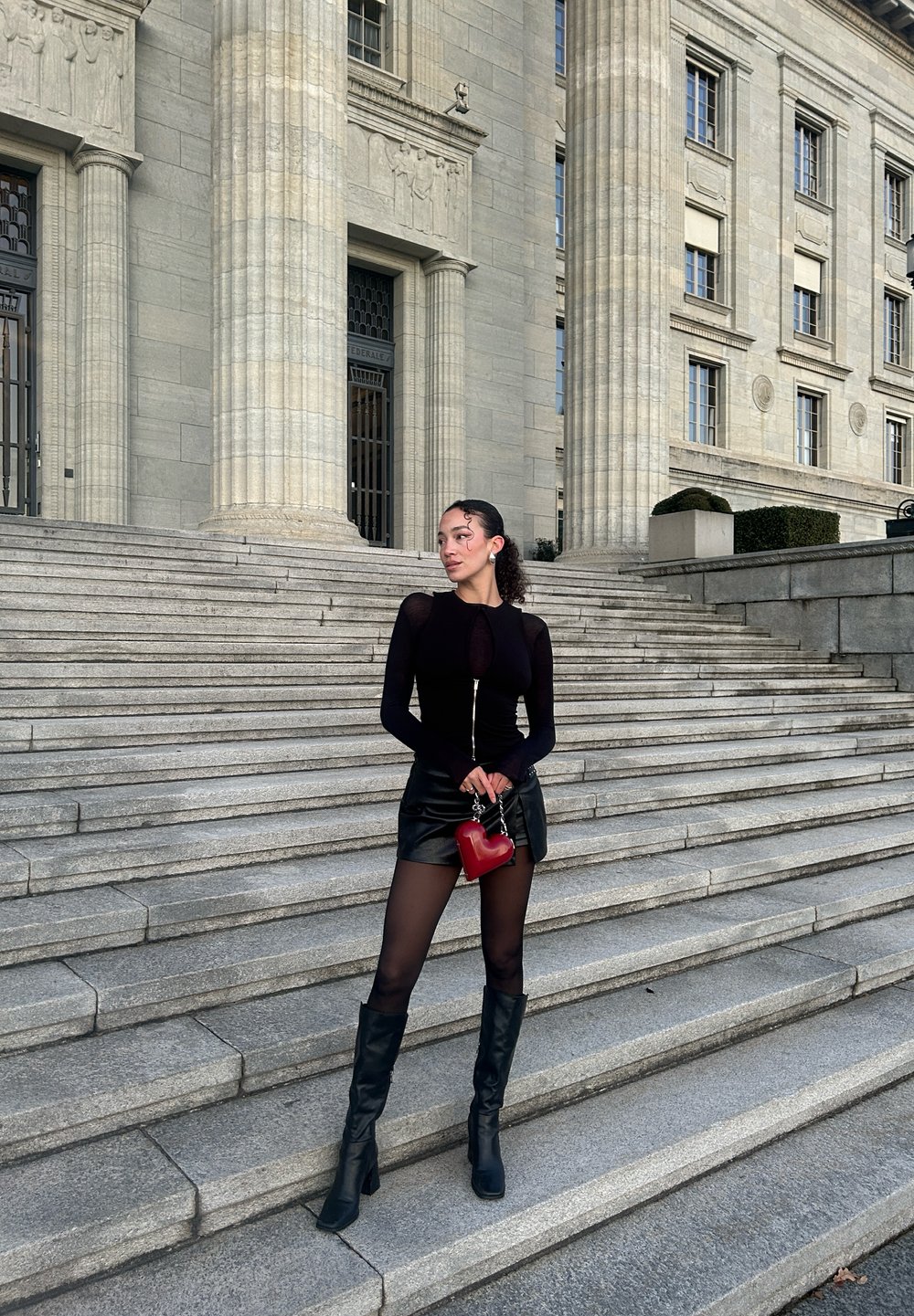 Črn bodysuit na zadrgo, v paru z mini črno krilo, prozornimi nogavicami in črnimi škornji do kolen. Drži rdečo torbico v obliki srca. Ozadje z kamenitimi stopnicami.