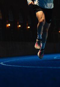 Jambes d'un coureur en plein saut sur une piste bleue la nuit, portant des chaussettes blanches et des chaussures de course avec des accents roses, avec des lumières extérieures tamisées en arrière-plan.